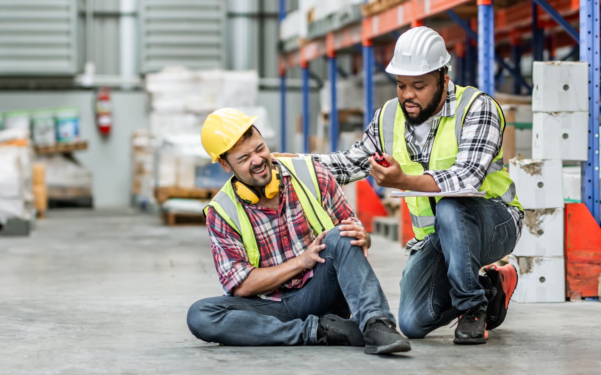 A coworker kneels beside an injured warehouse worker holding his knee